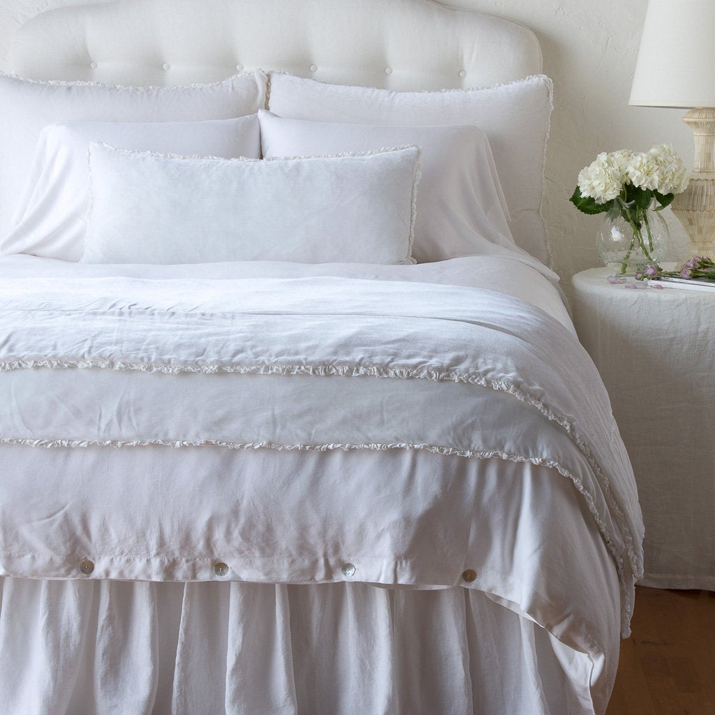 A white Carmen throw blanket with a petite charmeuse ruffle on a bed, with a nightstand and lamp beside it.
